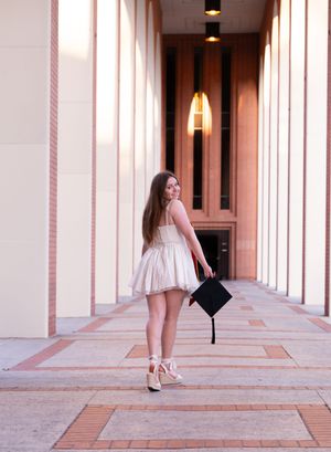 Graduate in a light pink dress holding a mortarboard while posing between architectural columns on campus.