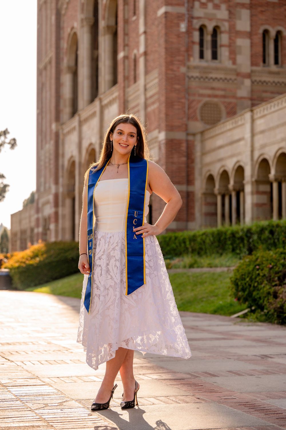 A graduate in a white lace dress and blue graduation stole stands on a sunny walkway by ornate brick architecture.