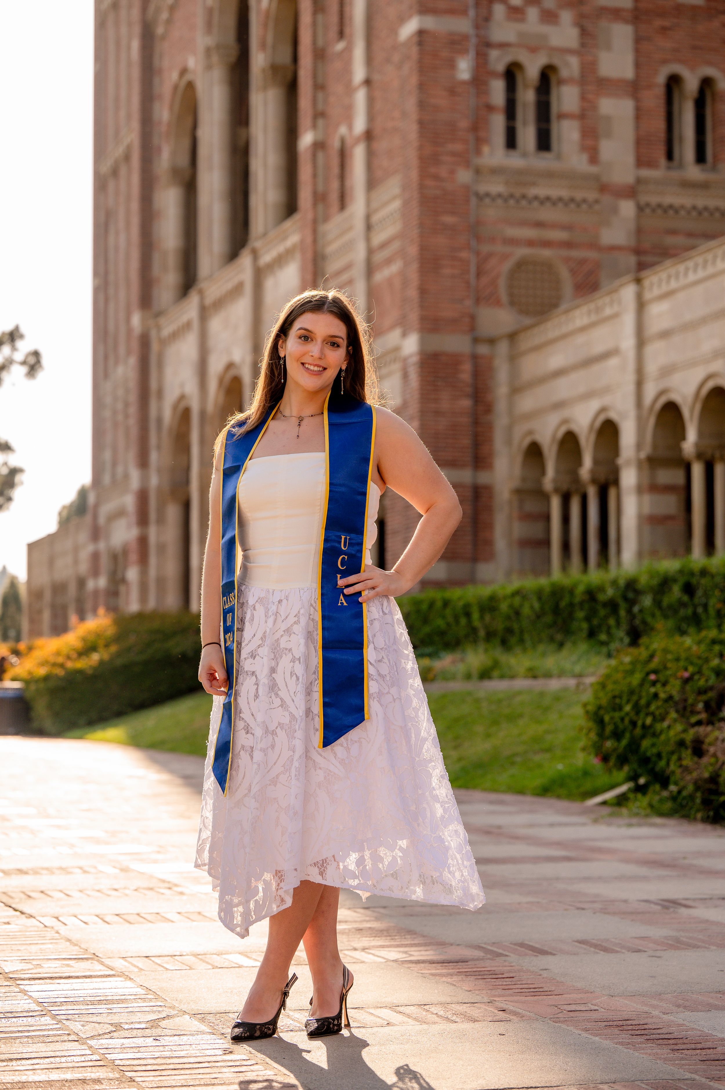 A graduate in a white lace dress and blue graduation stole stands on a sunny walkway by ornate brick architecture.