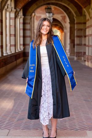 Graduate wearing black academic gown and bright blue stole poses in elegant lace dress under arched university hallway.