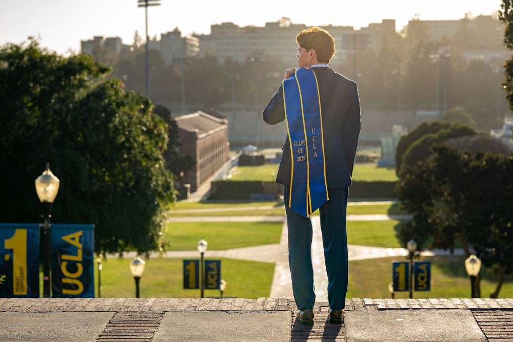 Graduate wearing blue sash and suit stands on steps overlooking campus during golden hour sunset.