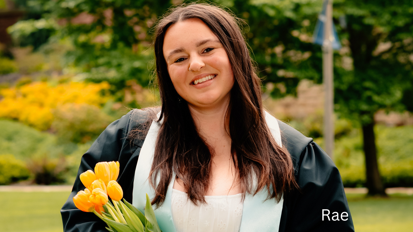 Graduate in black gown holding yellow tulips against green garden background.
