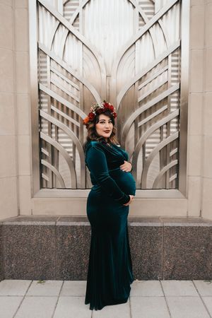 Expectant mother in elegant teal velvet gown poses against Art Deco architectural backdrop.