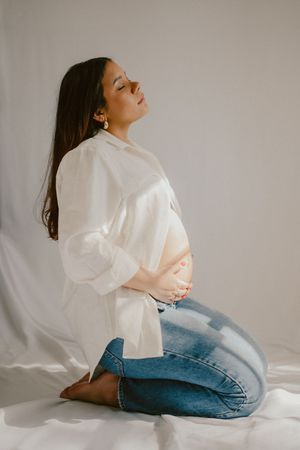 A pregnant woman in a white button down shirt and jeans poses serenely against a neutral background.