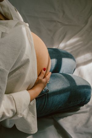 Close-up of a pregnant belly with hands gently resting on it, wearing white shirt and jeans.