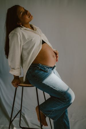 A stylish maternity photo of a woman in white shirt and jeans sitting on a stool.