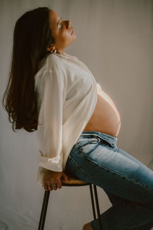 A profile shot of an expectant mother leaning back on a stool in casual attire.