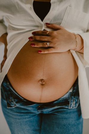 Close-up of a pregnant belly with red painted nails and jewelry visible against white shirt and jeans.
