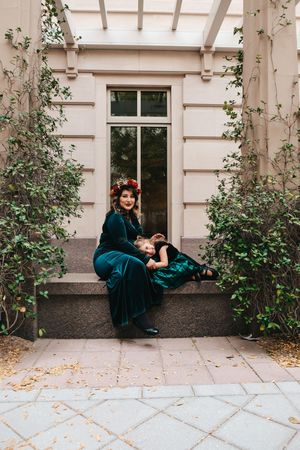 Person in emerald velvet dress sits on stone steps surrounded by climbing vines and architecture.