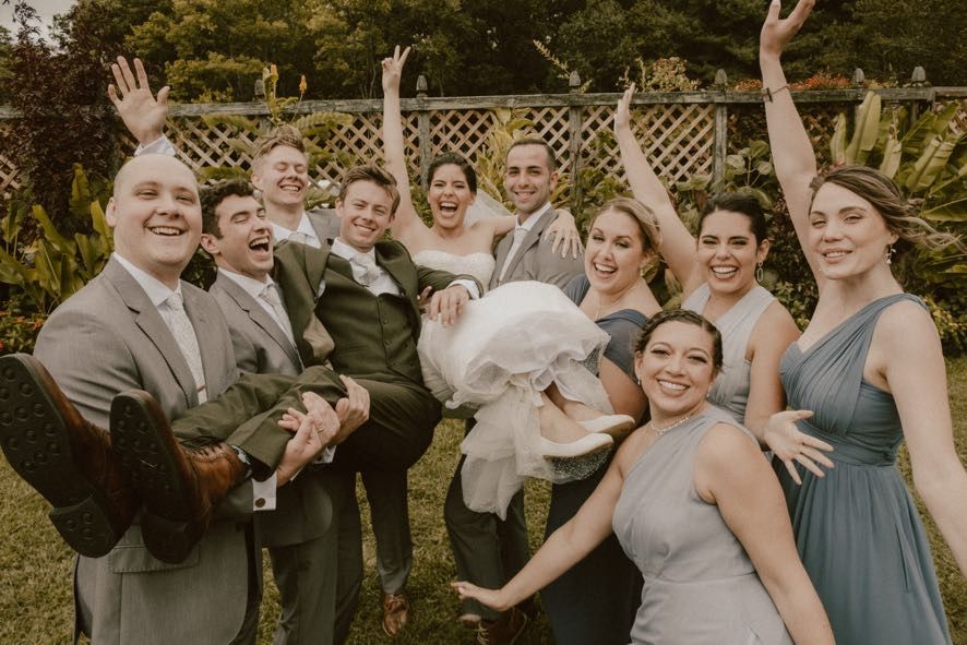 A joyful wedding party poses together in a garden setting wearing gray and blue formal attire.