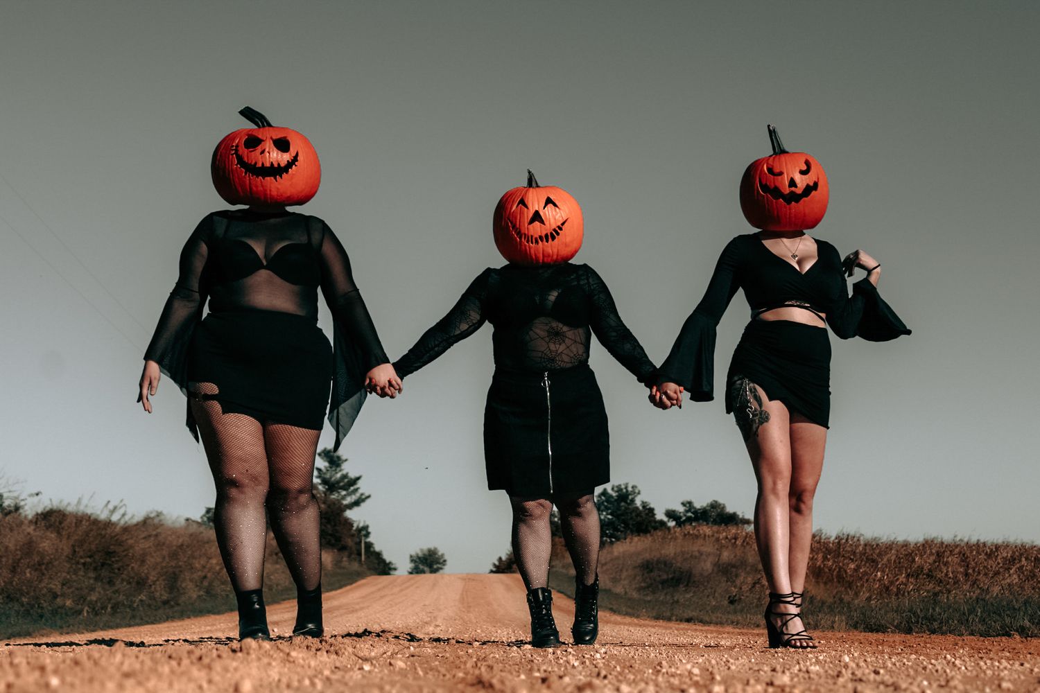 Three figures in black outfits wearing pumpkin heads hold hands walking on dirt path.