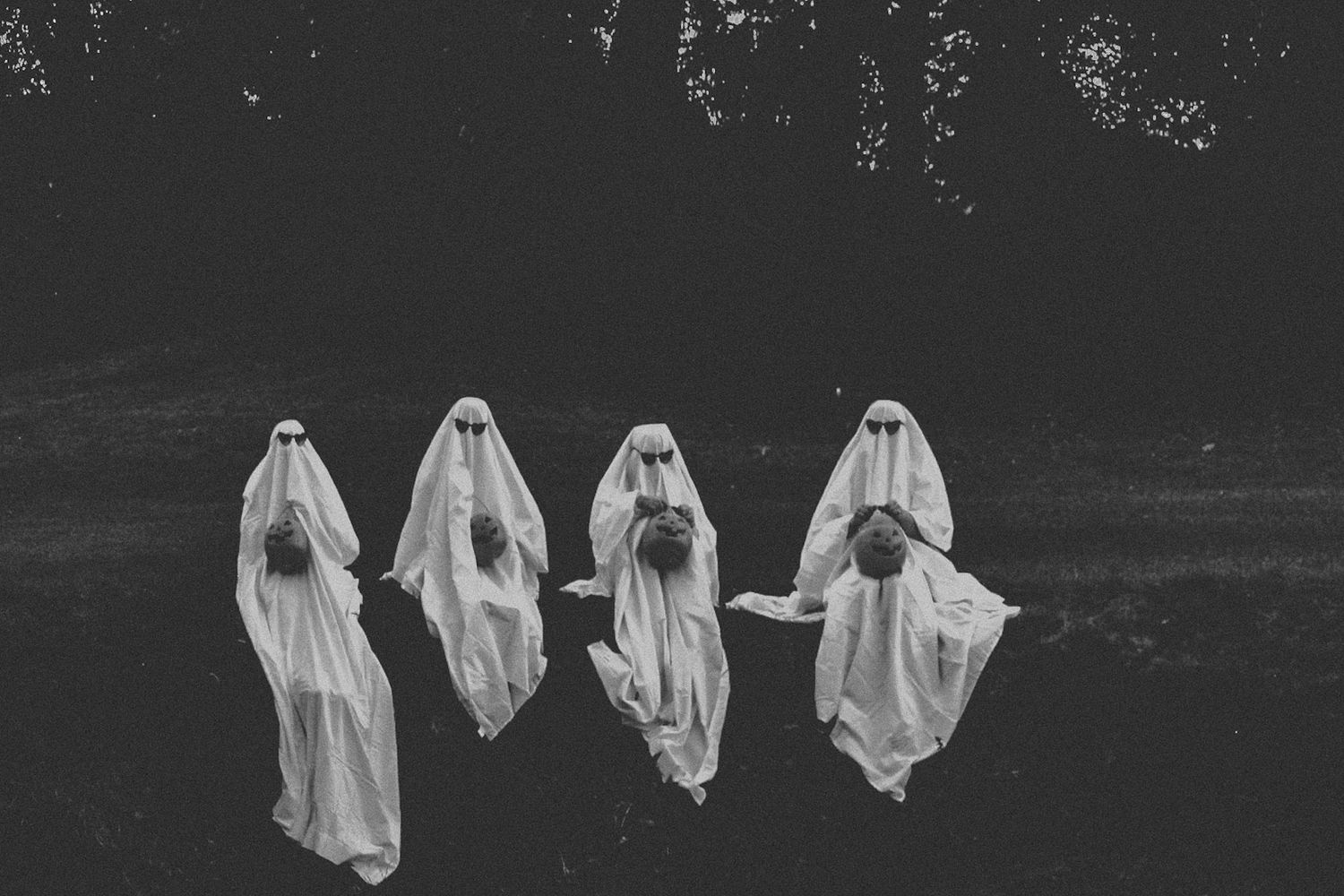 Four ghost figures in white sheets pose together in a spooky black and white forest setting.