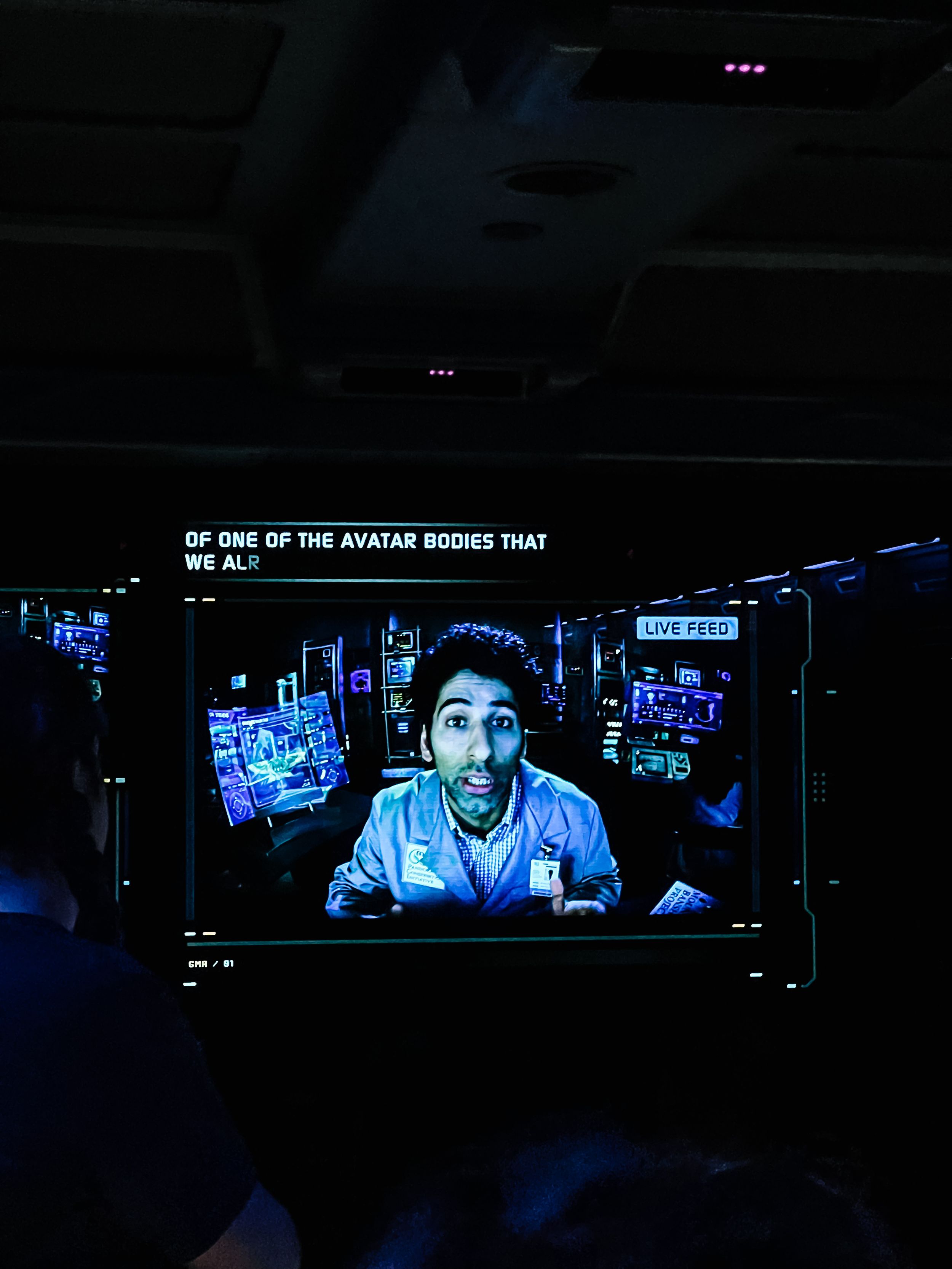 A dimly lit screen displaying someone speaking in a dark room with blue digital interfaces in the background.