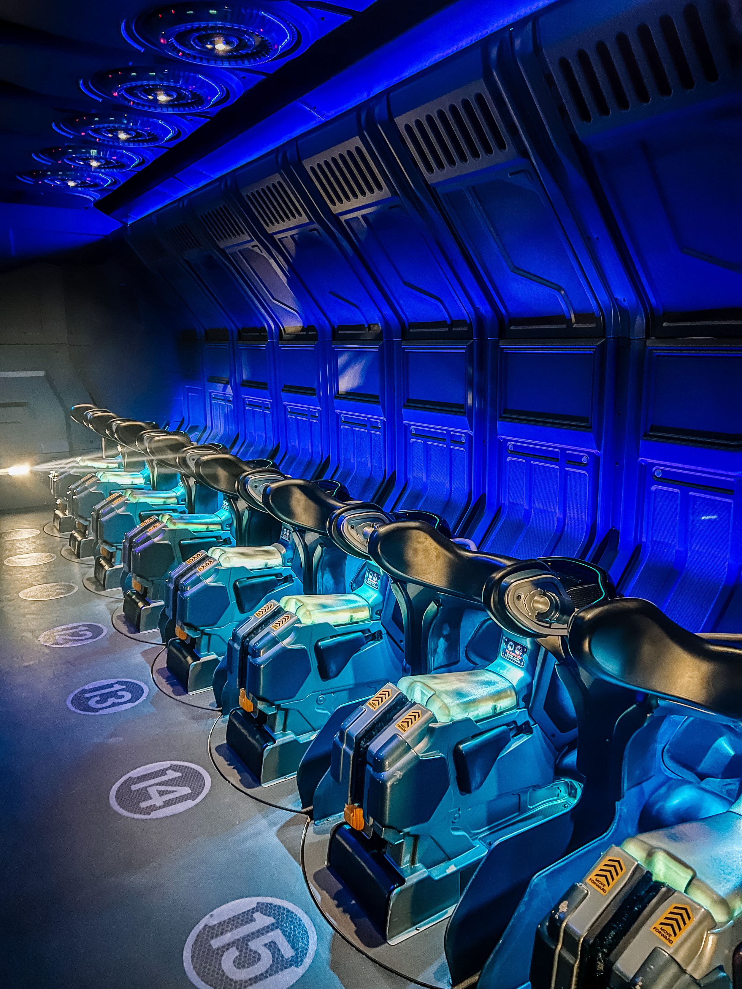 Illuminated blue and turquoise transport pods with technical displays line a sleek futuristic corridor at Space Mountain attraction.