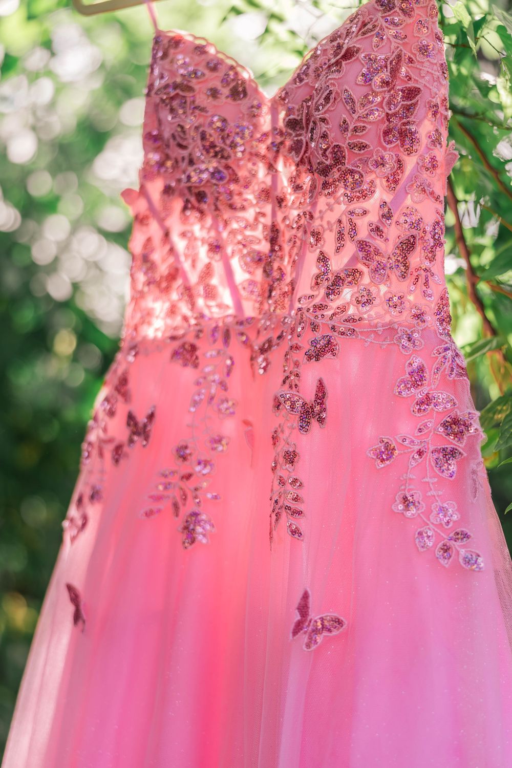 Pink sequined formal gown with butterfly details hanging outdoors against green foliage.