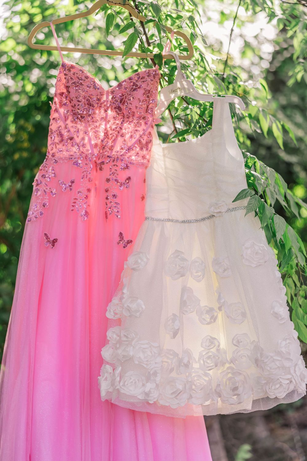 Pink and white formal dresses with butterfly embellishments displayed on hangers among tree branches.