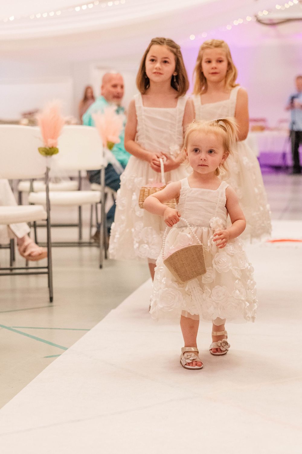 Young attendants in white dresses walking down aisle during wedding ceremony.