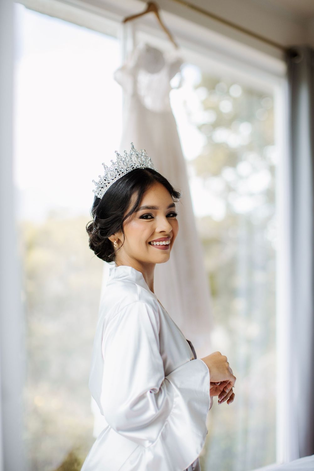 Bride in white robe poses by window with wedding dress hanging in background.