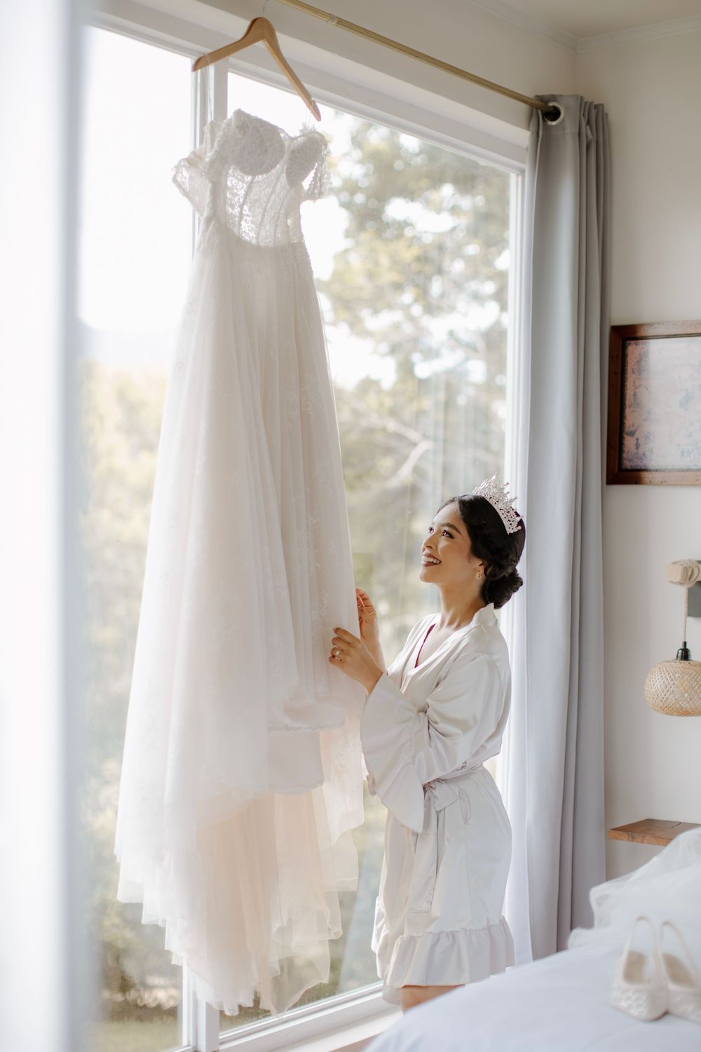 A white wedding gown hangs by a sunlit window as preparations begin for the special day.