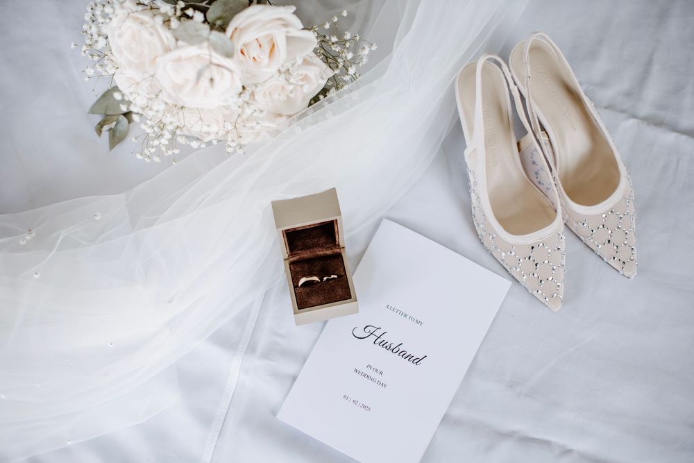 Wedding details including white heels, rings in a box, and delicate roses on white fabric.