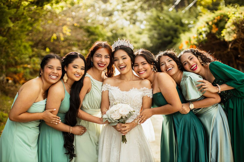 Bridesmaids in mint and emerald dresses gather joyfully outdoors for a wedding photo.