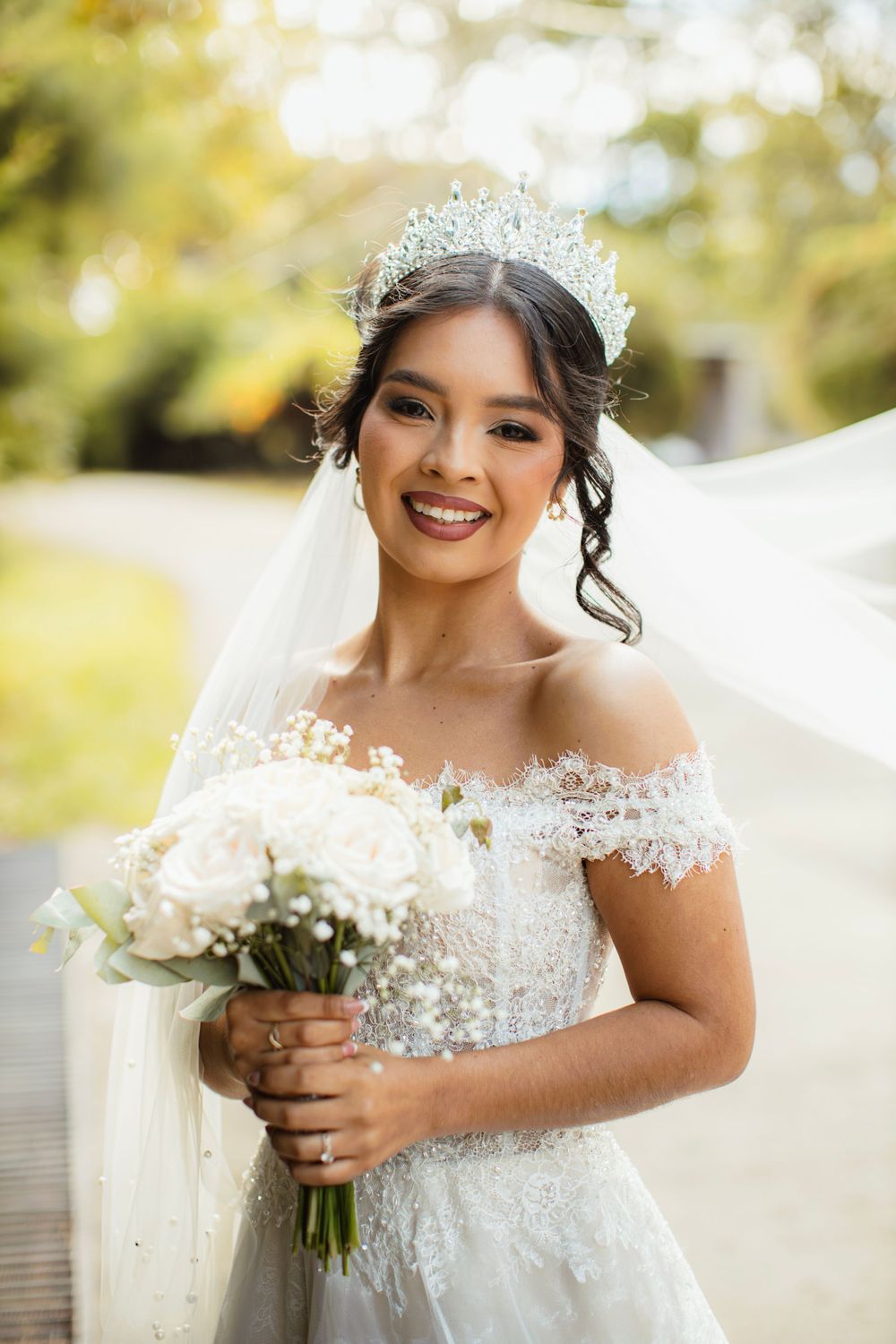 A radiant bride in a lace off-shoulder gown holds a white bouquet outdoors.