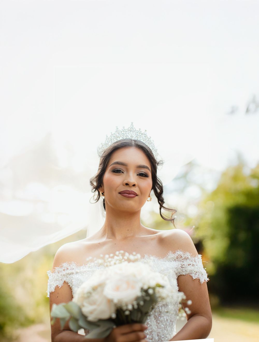 Bride in off-shoulder gown holds white bouquet while wearing sparkling tiara.
