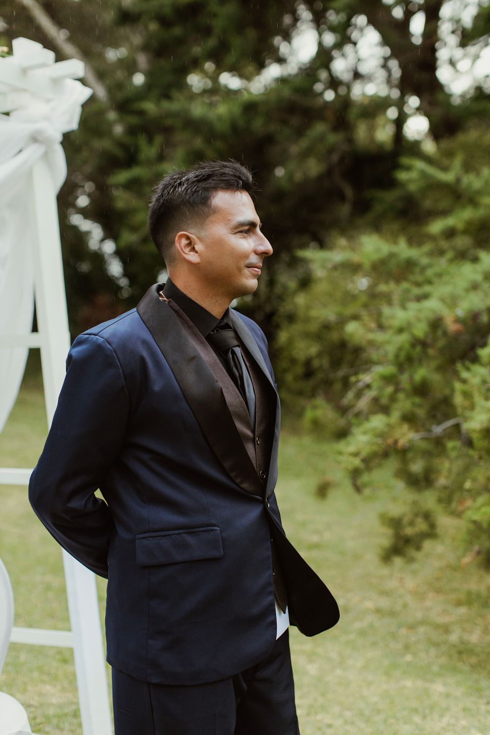 A formal portrait of a groom in a navy suit at an outdoor wedding ceremony.