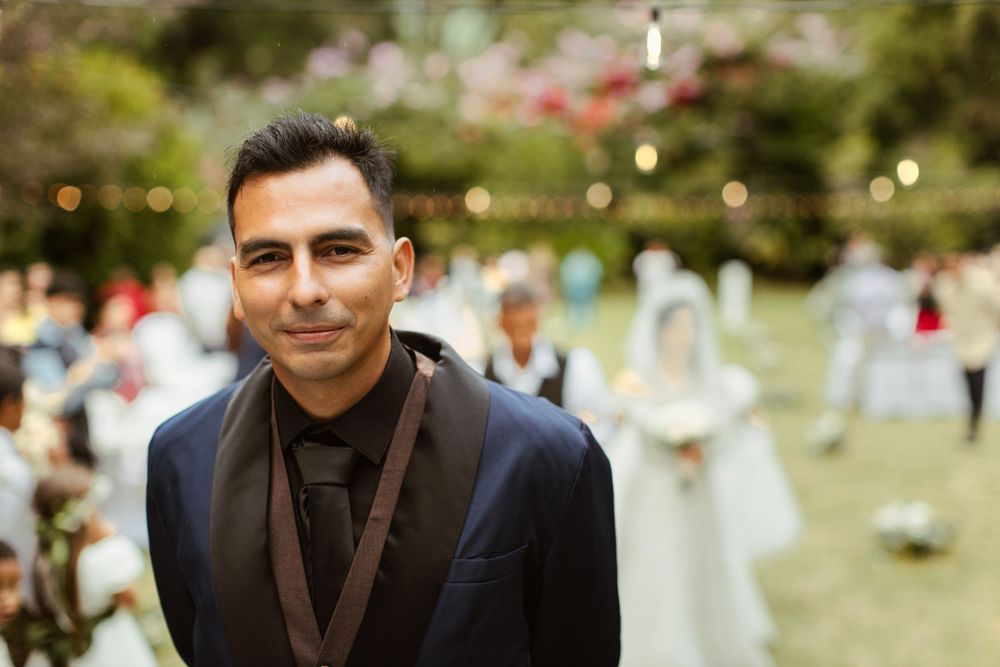 A groom in a dark suit smiles during an outdoor garden wedding celebration.