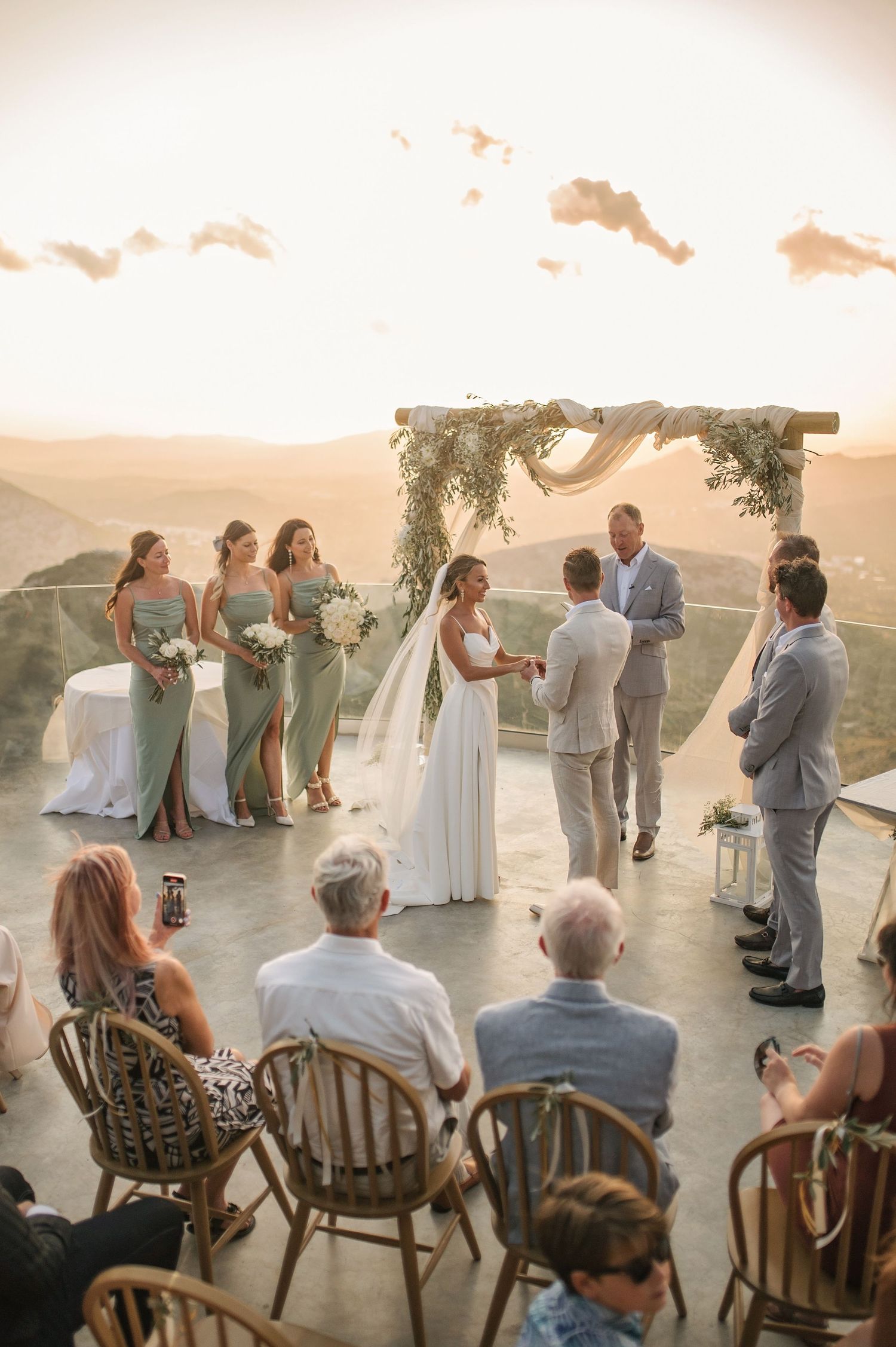 Intimate outdoor wedding ceremony on a hilltop at sunset with floral arch and rustic wooden chairs.