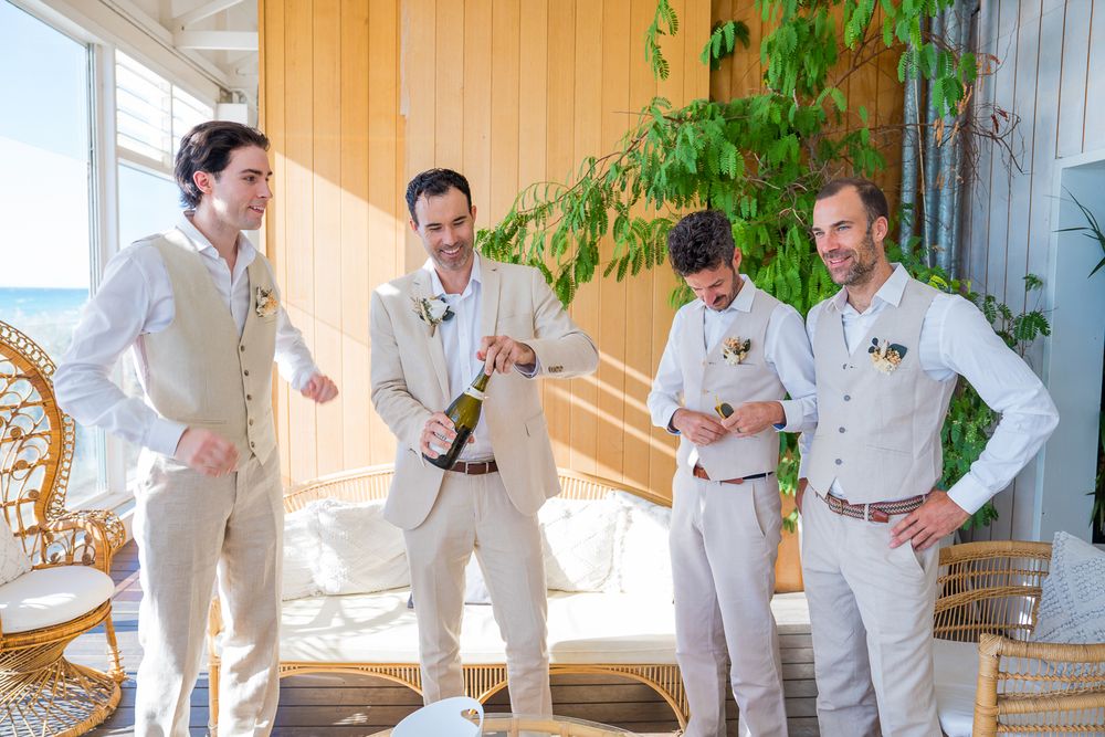 Four people in light-colored suits share a laugh while standing in a sunlit room with plants and wicker furniture.
