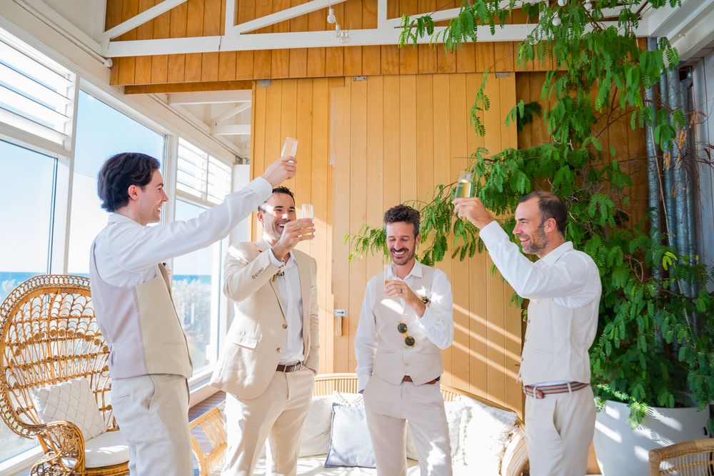 A group celebrates in a bright sunroom with wooden walls and wicker furniture, all dressed in light colored attire.