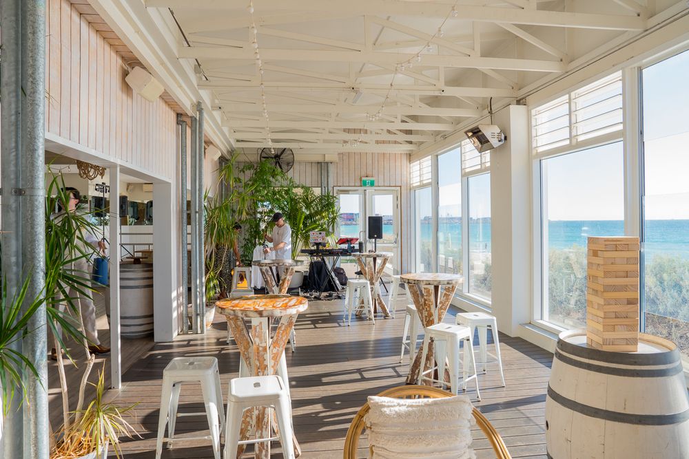 Bright coastal restaurant with white pillars, potted plants, and ocean views through large windows along the veranda.