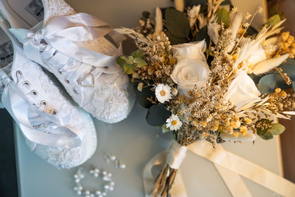 A detailed wedding styling flat lay featuring white lace shoes and a dried floral bouquet with golden wheat accents.