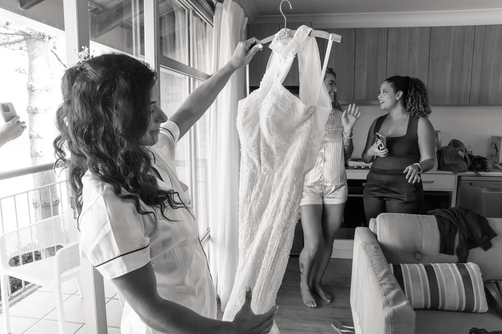 Black and white photo showing a wedding dress hanging while people prepare in a sunlit room.