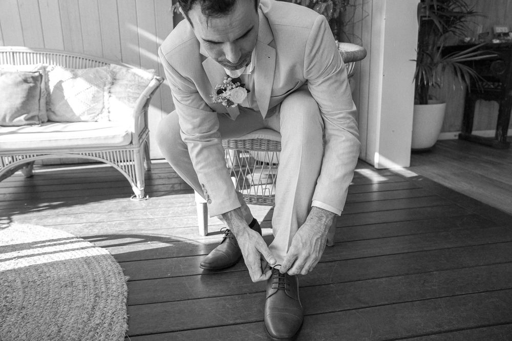 Black and white photo of someone in a light suit jacket sitting and tying their dress shoes on a wooden porch.