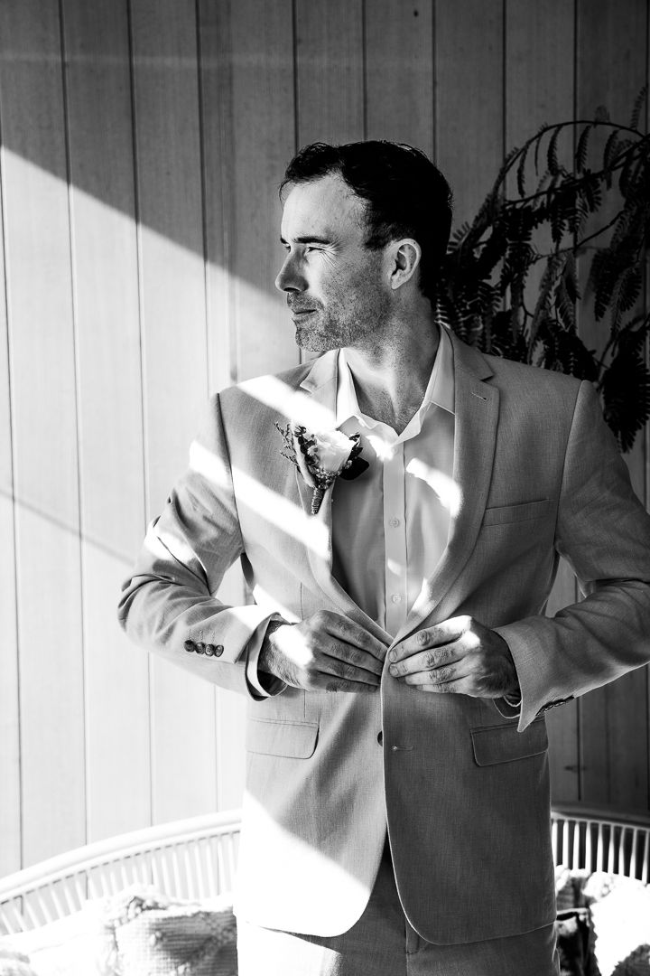 A black and white portrait of someone in a light colored suit jacket standing in dramatic side lighting.