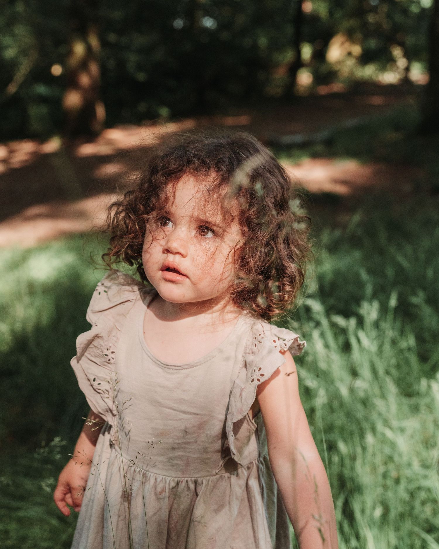 A curly-haired toddler wearing a light beige dress stands in dappled sunlight among grass and trees.