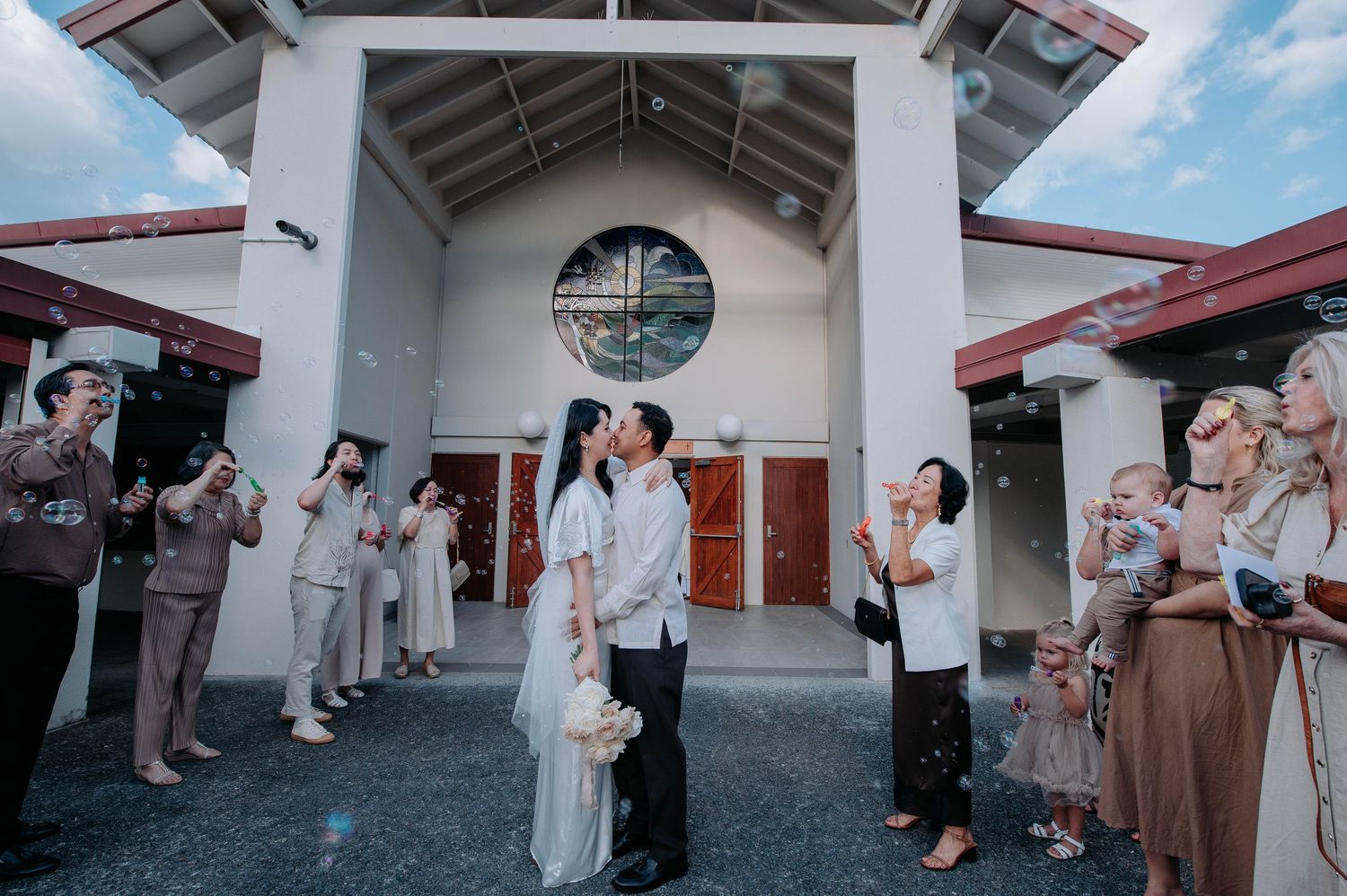 A couple shares a romantic moment outside a white chapel while guests celebrate with bubbles and photos.