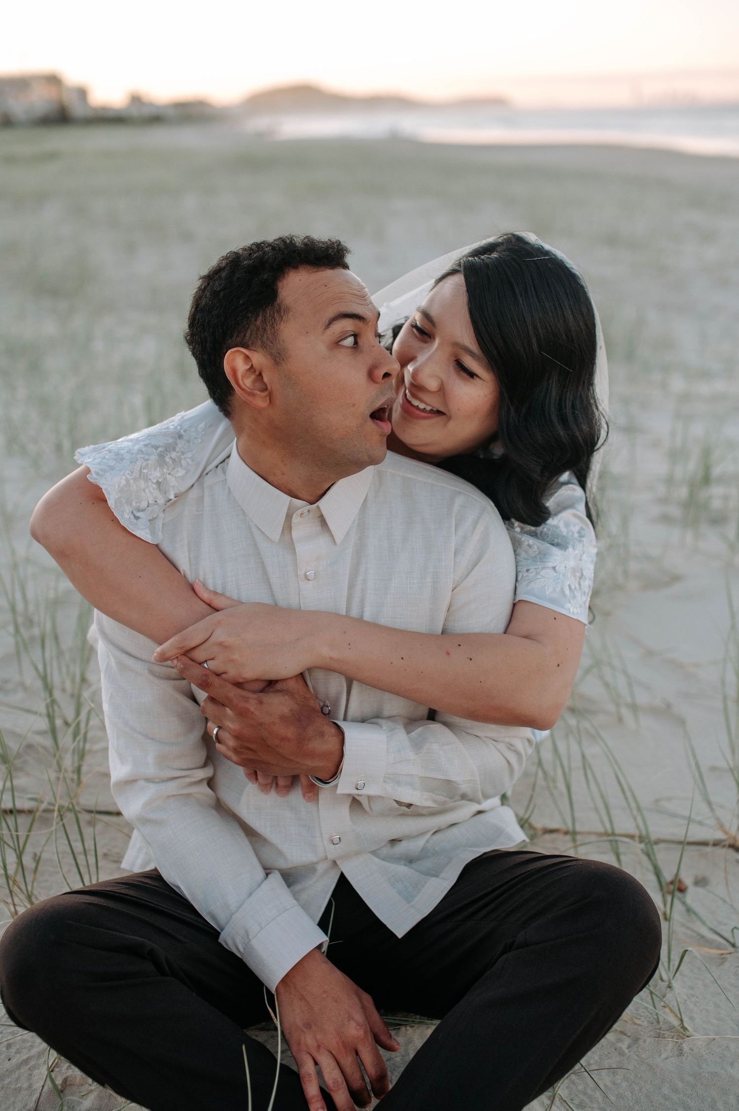 A loving couple embraces and laughs together on a sandy beach at sunset, both wearing white tops.