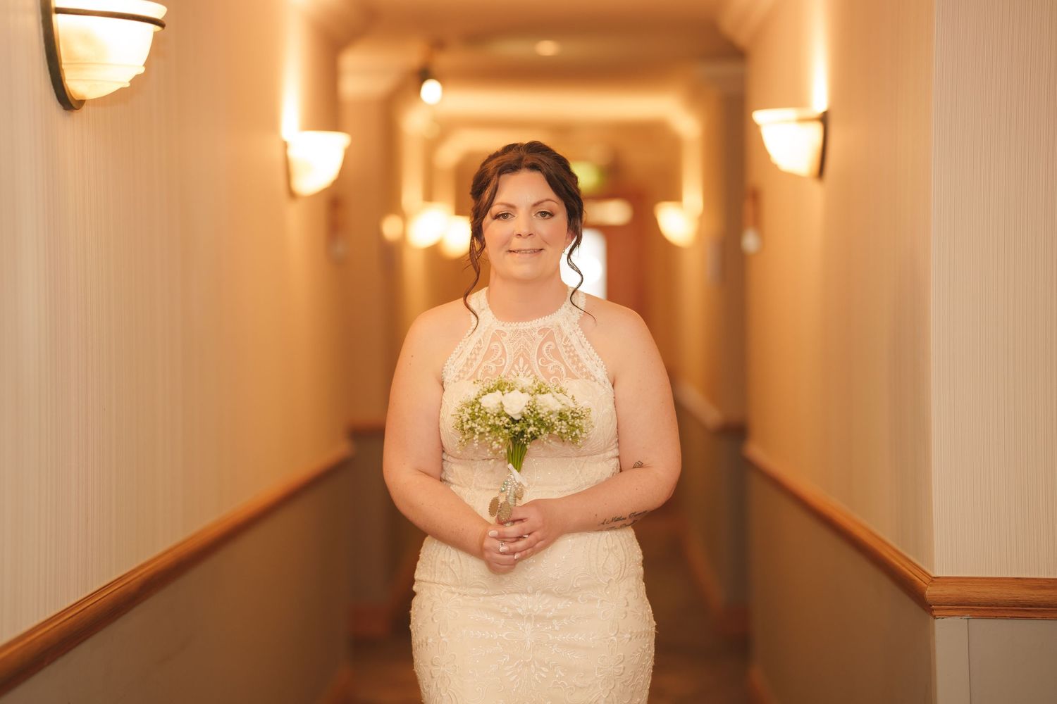 Bride holding bouquet in fitted lace gown while standing in warmly lit hotel hallway.