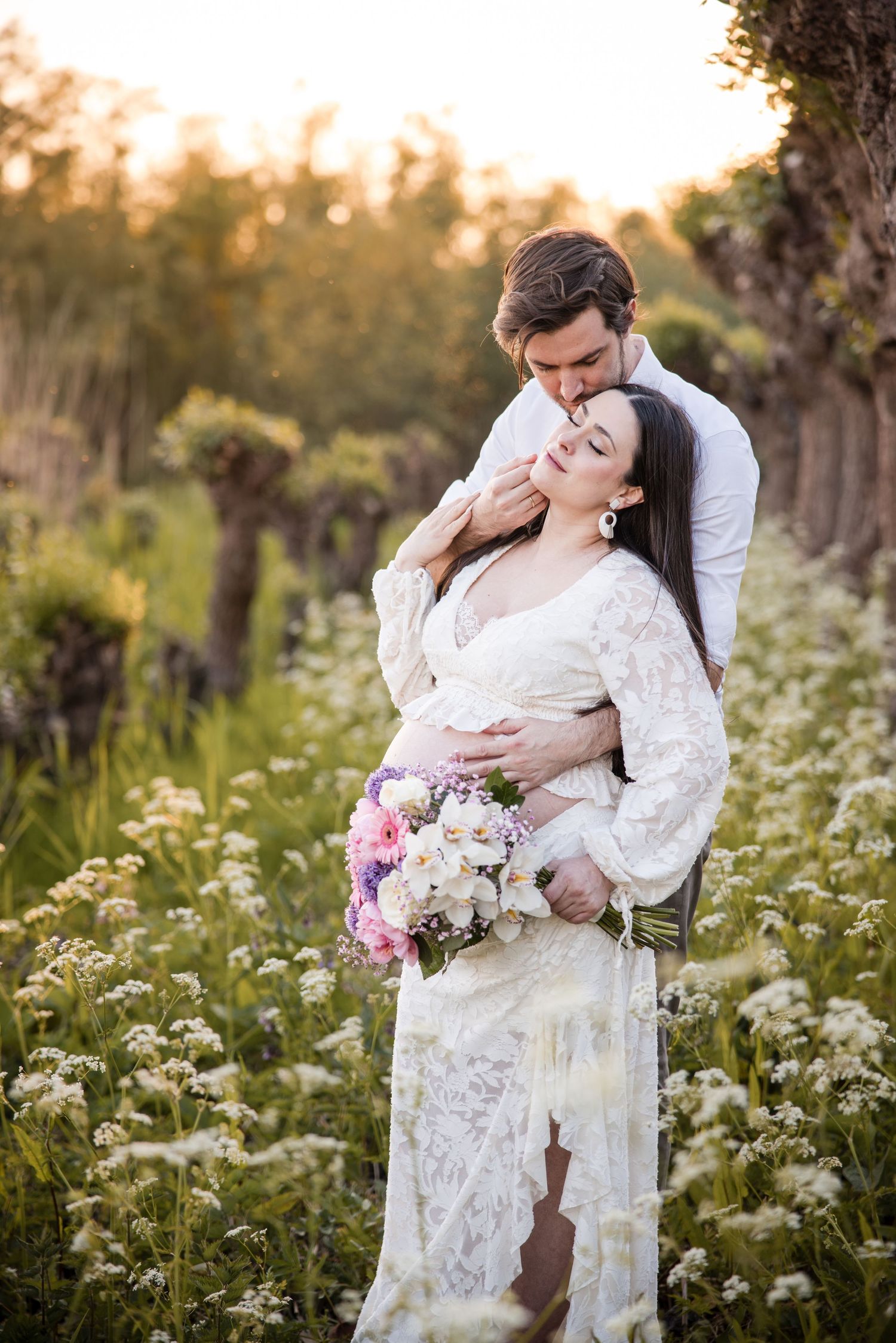 Romantic couple embraces in a dreamy wildflower meadow at sunset wearing white lace and holding a floral bouquet.