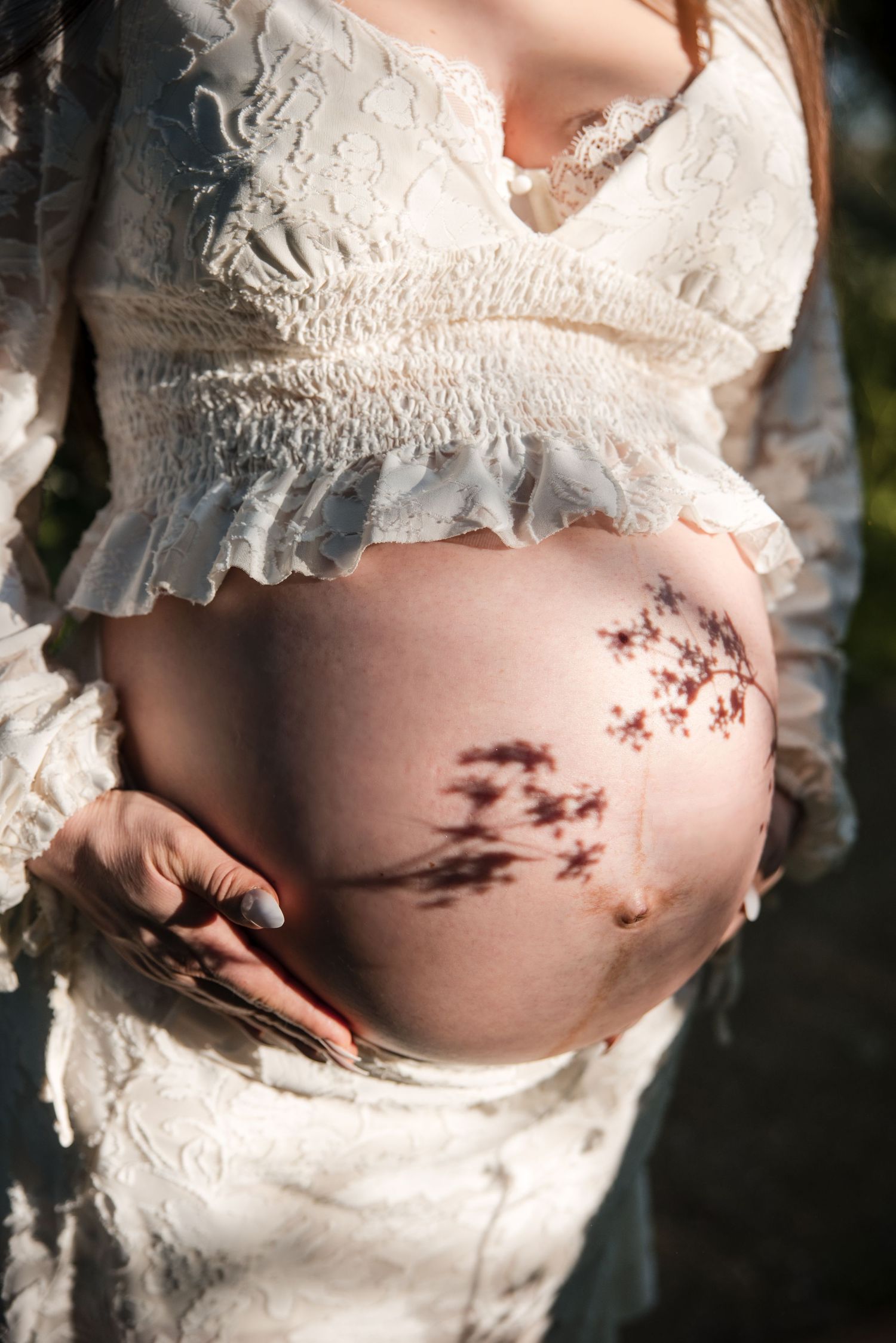 A pregnant belly with delicate lace dress details and soft shadows of wildflowers leaves cast across the skin in natural light.