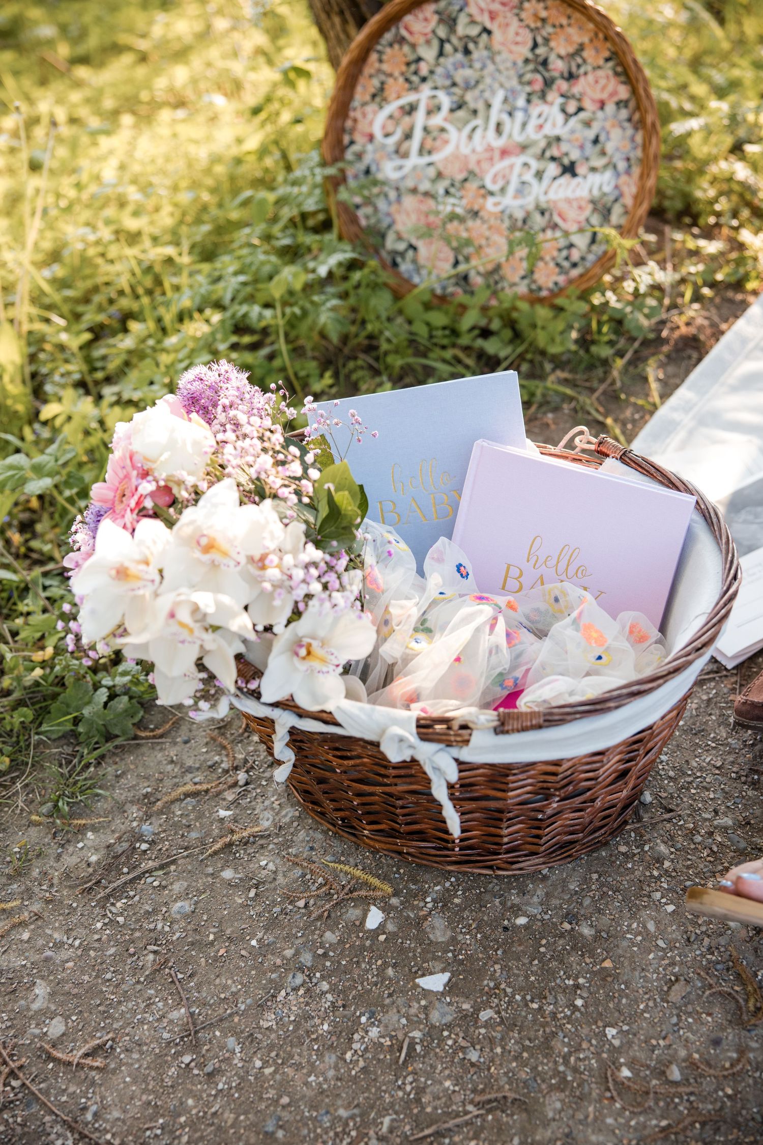 A wicker basket filled with baby items and floral arrangements sits beside a rustic decorative sign in a garden setting.