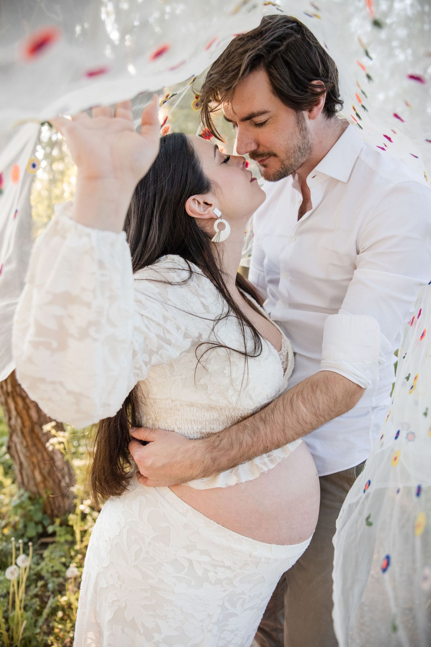 A romantic couple embraces and shares tender moments under a veil with colorful flowers around them in a dreamy outdoor setting.