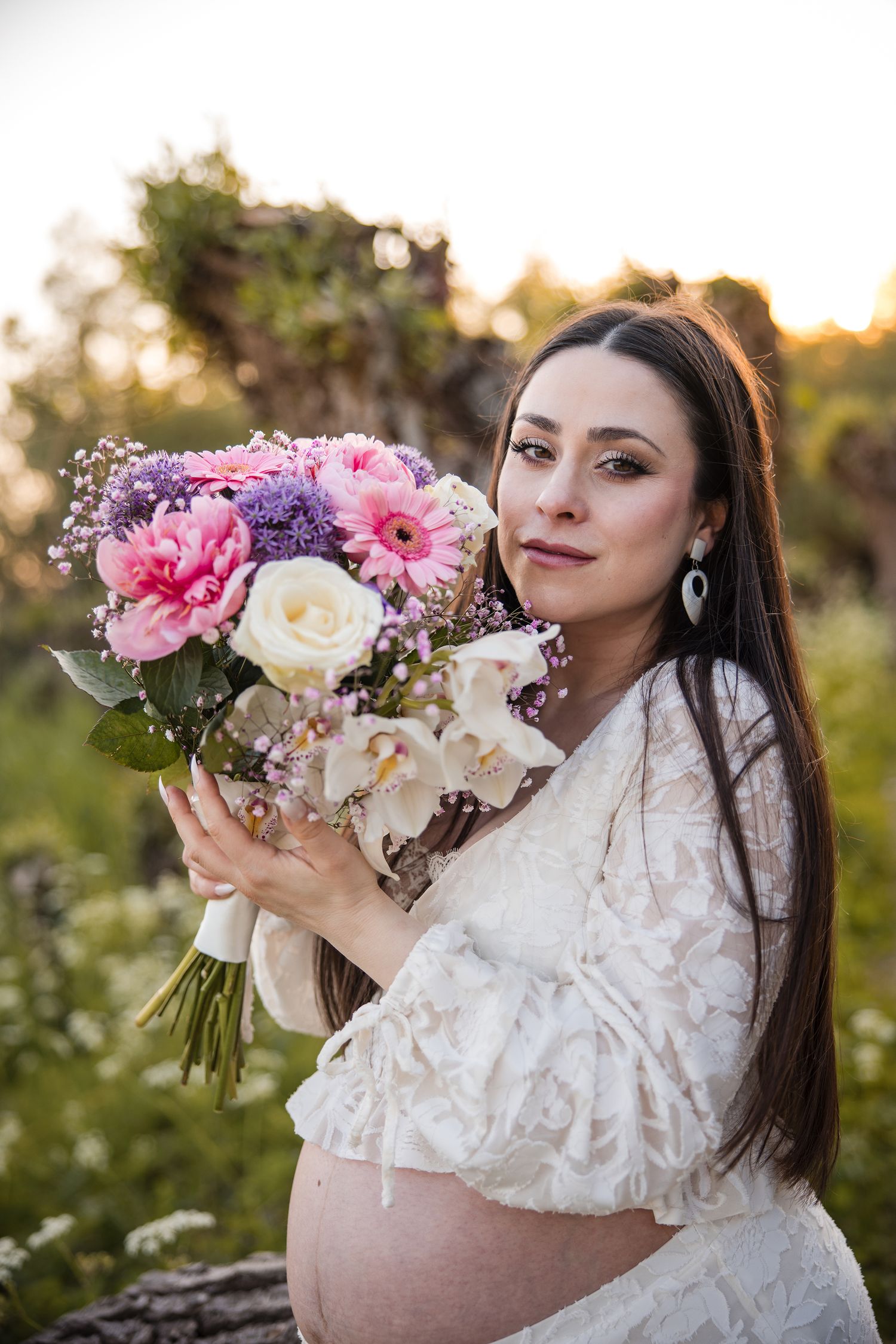 A pregnant woman in a white lace dress holds a vibrant bouquet of pink and white flowers during a sunset photoshoot.