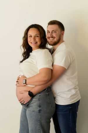 Couple in white t-shirts embracing against white wall in maternity photoshoot.