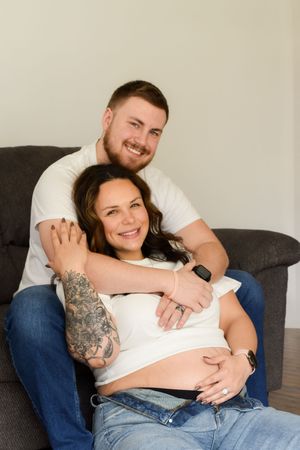 Couple relaxes together on gray couch as they lovingly embrace while expecting a baby.