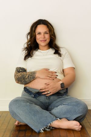 Woman in white t-shirt and distressed jeans sits cross-legged on wooden floor with visible tattoo sleeve.