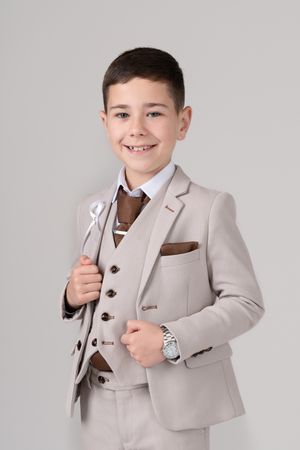 Young child wearing a formal beige three-piece suit with brown tie and pocket square poses against gray background.